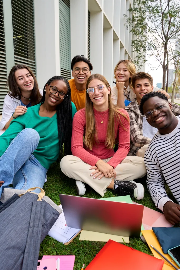 Group of diverse students sitting outdoors with notebooks and a laptop, smiling together.